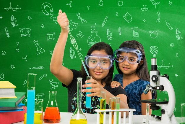 Dos pequeñas y lindas colegialas indias o asiáticas experimentando o estudiando ciencias en laboratorio, sobre fondo de pizarra verde con garabatos educativos