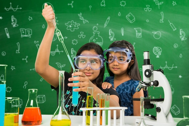 Foto dos pequeñas y lindas colegialas indias o asiáticas experimentando o estudiando ciencias en laboratorio, sobre fondo de pizarra verde con garabatos educativos