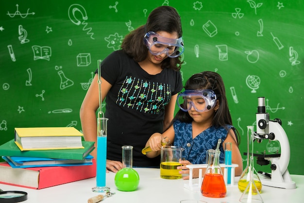 Dos pequeñas y lindas colegialas indias o asiáticas experimentando o estudiando ciencias en laboratorio, sobre fondo de pizarra verde con garabatos educativos