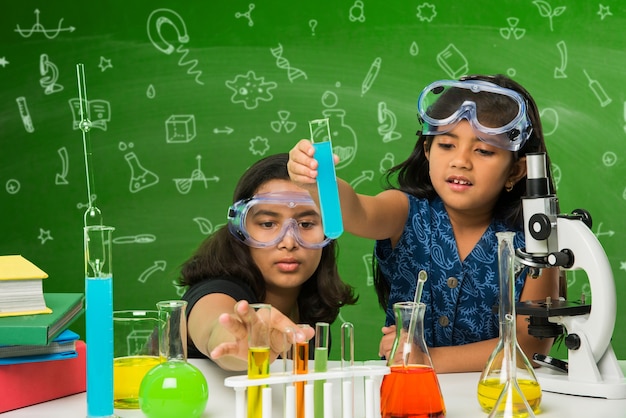 Dos pequeñas y lindas colegialas indias o asiáticas experimentando o estudiando ciencias en laboratorio, sobre fondo de pizarra verde con garabatos educativos