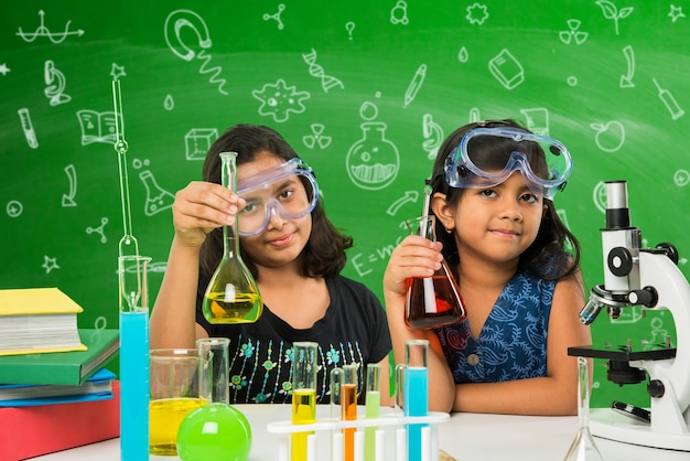 Dos pequeñas y lindas colegialas indias o asiáticas experimentando o estudiando ciencias en laboratorio, sobre fondo de pizarra verde con garabatos educativos