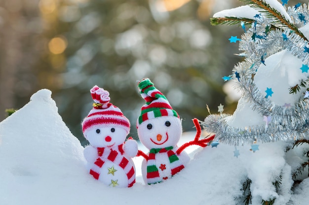 Dos pequeños juguetes bebé muñeco de nieve con sombreros de punto y bufandas en nieve profunda cerca de la rama de pino