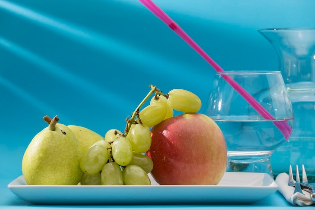 Dos peras, uvas y una manzana en una bandeja blanca detrás de un vaso con pajitas y una botella de agua.