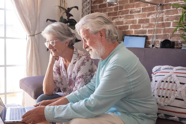 Dos personas mayores modernas maduras caucásicas sentadas en un sofá en casa usando una computadora portátil Pareja de ancianos de pelo gris relajándose y sonriendo juntos mirando la misma computadora portátil Fondo de pared de ladrillo