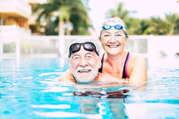 Dos personas mayores o personas maduras abrazados juntos en el agua azul de la piscina - hombre y mujer activos haciendo ejercicio juntos - verano y divirtiéndose