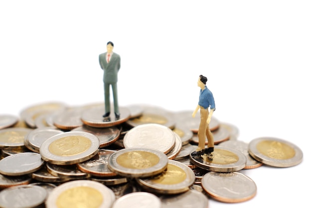 Dos personas en miniatura caminando y de pie sobre un montón de monedas de baht tailandés aislado sobre fondo blanco.