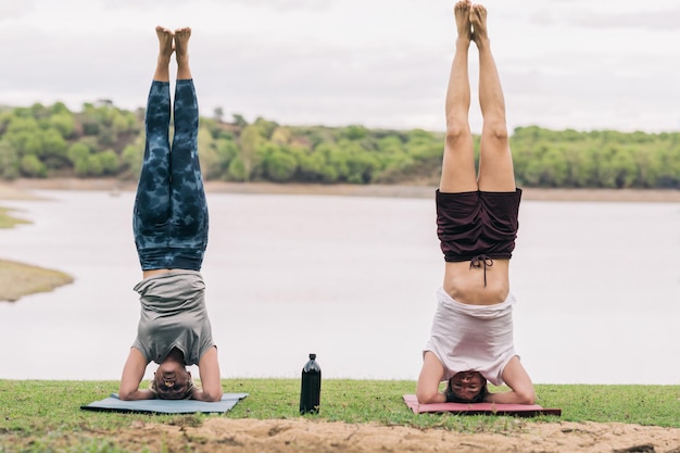 Dos personas practicando yoga en posición inversa de la cabeza al suelo junto a un lago en un parque.