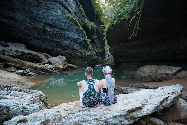 Dos personas se sientan en una gran roca y miran la gruta, las rocas y el río de la montaña, el lago entre las rocas, el bosque tropical