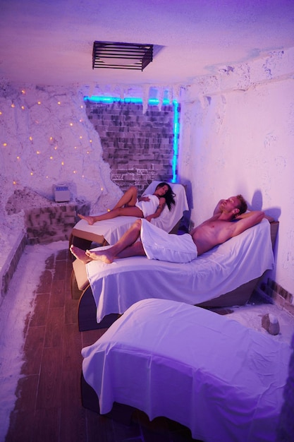 Dos personas en toallas blancas acostadas en camas interiores en una habitación con decoración violeta