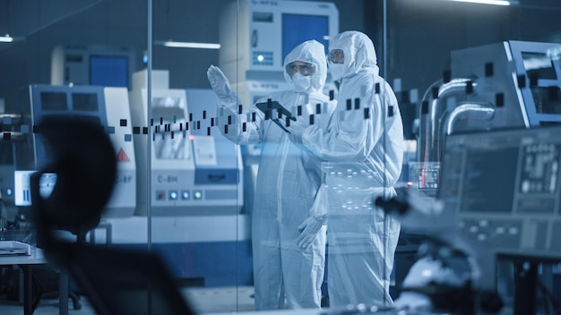 Dos personas con trajes protectores se paran frente a una pared de vidrio con un equipo de laboratorio al fondo.