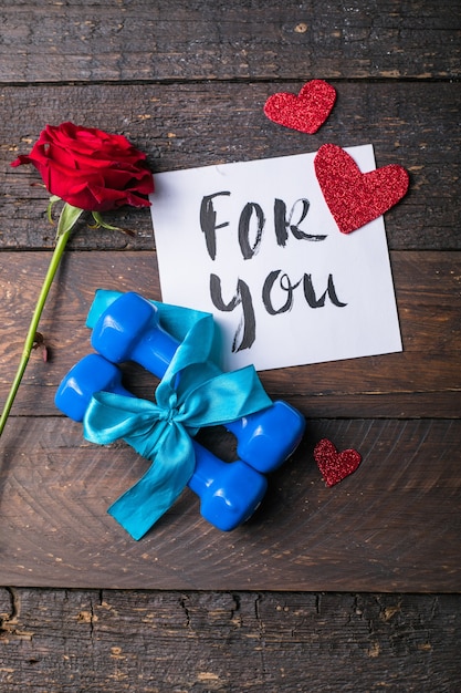 Dos pesas azules con un arco de regalo y una etiqueta de papel en blanco, corazón sobre tablas de madera, concepto de vacaciones deportivas