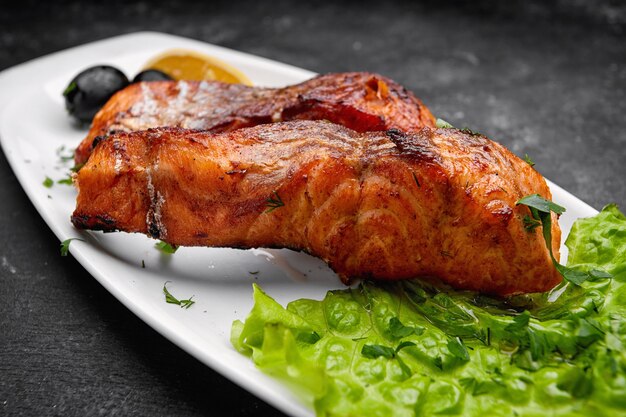 Dos piezas de salmón a la parrilla al horno sobre una placa blanca con lechuga, aceitunas y limón sobre una mesa oscura