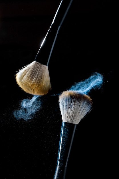 Dos pinceles para maquillaje con sombras de maquillaje azul en movimiento sobre un fondo negro.