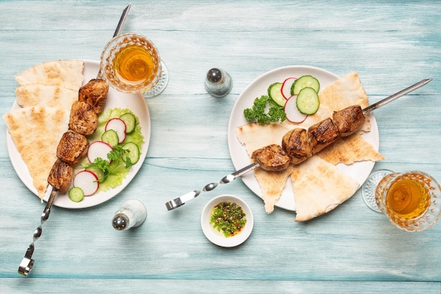 Dos porciones de apetitoso shish kebab casero, pepinos en rodajas y rábanos, copas de vino con vista superior sobre un fondo de madera azul.