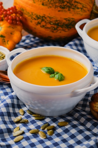 Dos porciones de sopa de calabaza casera con crema en un plato de caldo blanco sobre una servilleta a cuadros azul, picatostes, semillas de calabaza y albahaca sobre fondo de madera rústica, enfoque selectivo