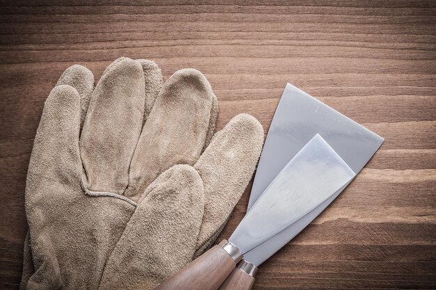 Dos raspadores de pintura y guantes de seguridad sobre tablero de madera.