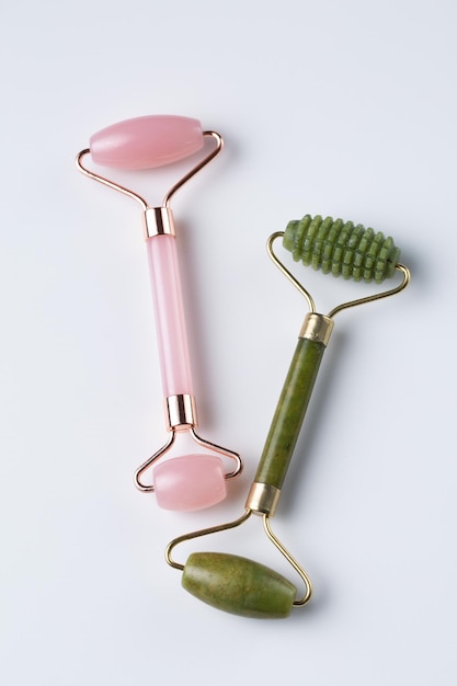 Dos rodillos faciales cosméticos de cristal de cuarzo rosa y jade aislados en fondo blanco vista superior