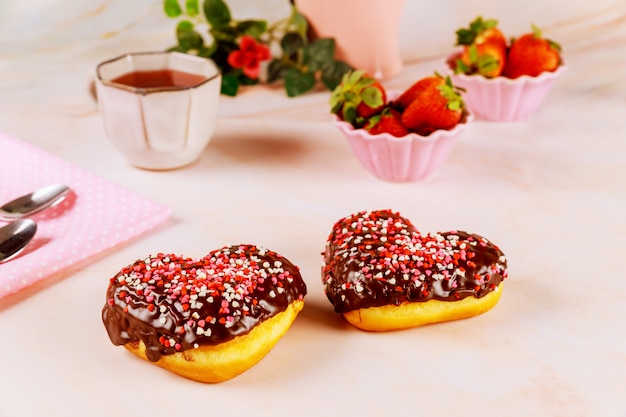 Dos rosquillas en forma de corazón con glaseado de chocolate y rosa, rocía rojo sobre una placa con fresa.