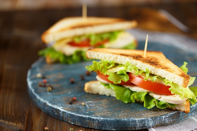 Dos sabroso sándwich con pollo, tomate, lechuga, queso en un plato de madera sobre un fondo oscuro