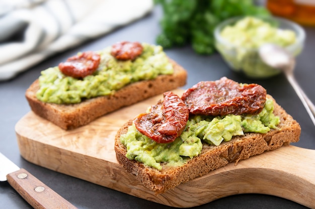 Dos sandwiches de aguacate. Pan de centeno con guakomole, pasta de aguacate y tomates secos, sobre tabla para cortar madera. Tostada de aguacate.