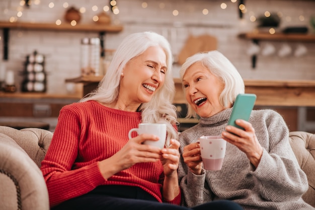 Dos señoras lindas agradables de pelo gris sentado en el sofá y riendo