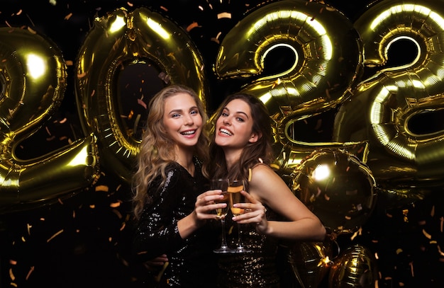 Dos señoritas bebiendo champán. Imagen de chicas con globos aislados sobre fondo negro, divirtiéndose en la fiesta de Navidad o Nochevieja 2022.