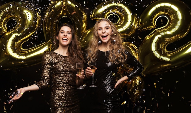 Dos señoritas bebiendo champán. Imagen de chicas con globos aislados sobre fondo negro, divirtiéndose en la fiesta de Navidad o Nochevieja 2022.