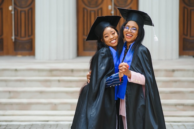 Dos señoritas en trajes de graduación posando a la cámara en el campus universitario, sosteniendo diplomas, riendo y abrazándose, teniendo feliz día de graduación, retrato de portarretrato