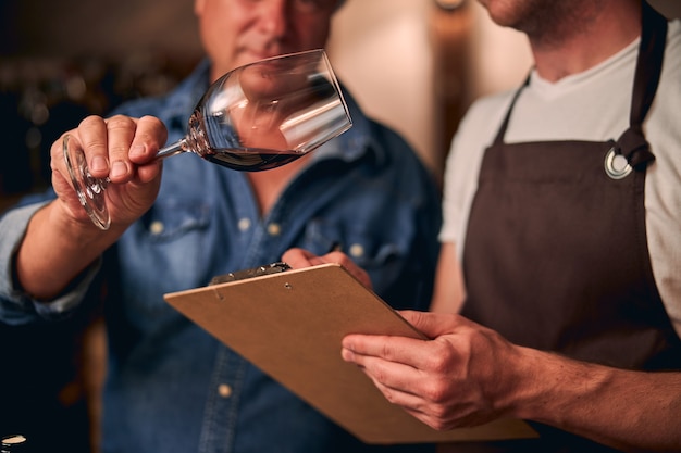 Dos sommeliers no reconocidos en una bodega tomando notas en un portapapeles mientras evalúan el vino tinto en una copa