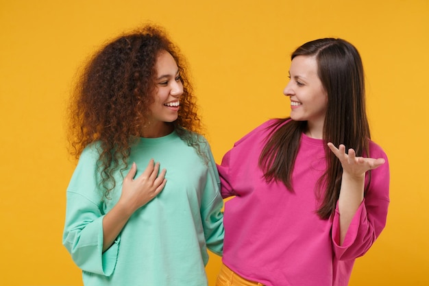 Dos sonrientes y alegres amigas europeas y afroamericanas vestidas de verde rosa posando aisladas en un fondo amarillo. Concepto de estilo de vida de las personas. Simulacros de espacio de copia. Mirando el uno al otro.