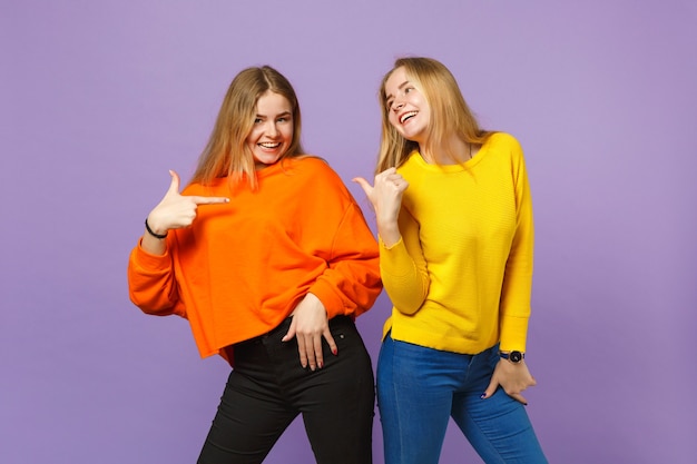 Dos sonrientes jóvenes rubias gemelas hermanas niñas en ropa de colores vivos señalando con el dedo el uno al otro aislado en la pared azul violeta pastel. Concepto de estilo de vida familiar de personas.