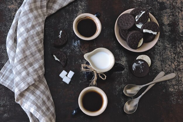 Dos tazas de café expreso, una jarra de leche y galletas de chispas de chocolate con un relleno blanco.