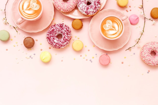 Dos tazas de café con leche, deliciosas rosquillas rosas con espolvorear y coloridos macarons brillantes