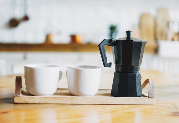 Dos tazas de cerámica blanca y una cafetera de géiser en una bandeja de madera en la mesa de la cocina