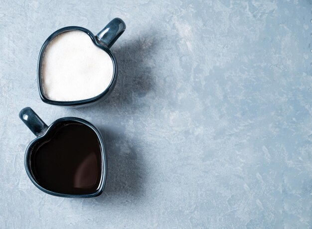 Dos tazas en forma de corazones con café aromático recién hecho con leche y espresso sobre fondo azul El concepto de bebidas opuestas Vista superior y espacio para copiar