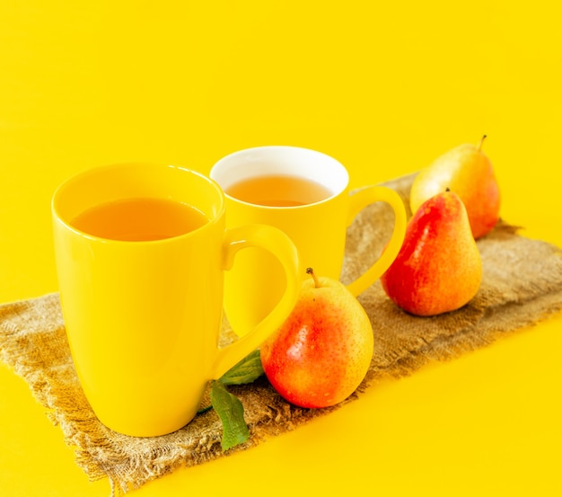 Dos tazas de jugo de pera y peras maduras sobre tela de arpillera y fondo amarillo.