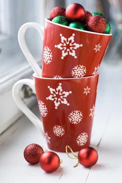 Dos tazas rojas con copos de nieve sobre fondo blanco de madera. Bolas de Navidad rojas y verdes en tazas.
