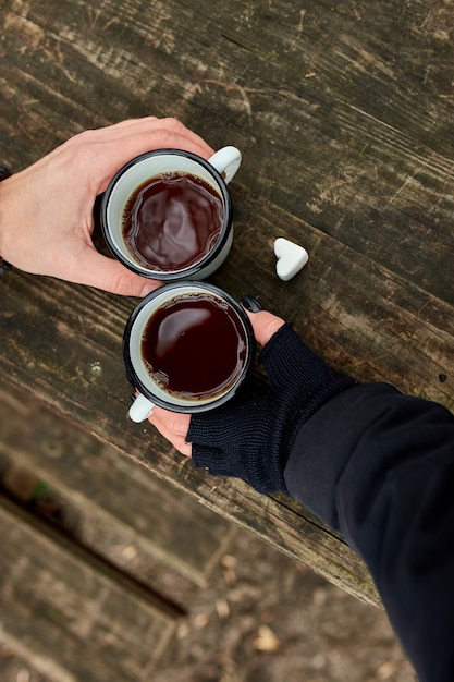 Dos tazas de té en la naturaleza en manos de la pareja en guantes negros sobre superficie de madera