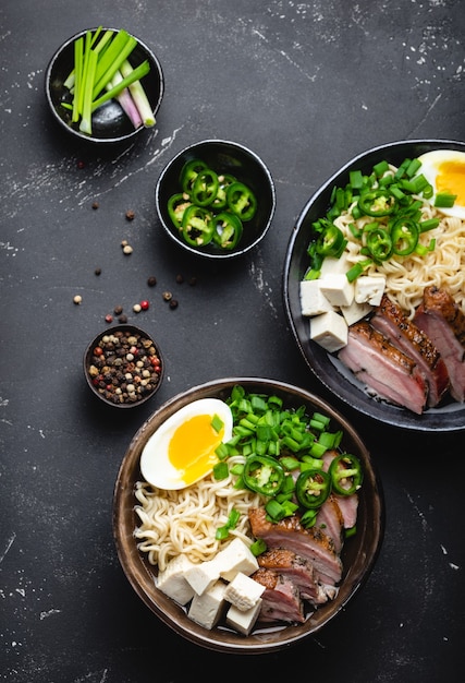 Dos tazones de ramen de sopa de fideos asiáticos sabrosos con caldo de carne, tofu, cerdo, huevo con yema sobre fondo de hormigón rústico negro, de cerca, vista superior. Sabrosa sopa de ramen japonesa caliente para cenar al estilo asiático