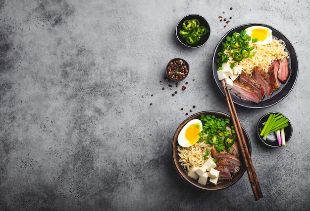 Dos tazones de ramen de sopa de fideos asiáticos sabrosos con caldo, tofu, cerdo, huevo sobre fondo de hormigón rústico gris, espacio para texto, primer plano, vista superior. Sabrosa sopa de ramen japonesa caliente para cenar con espacio de copia