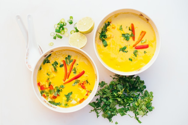 Dos tazones de sopa de curry de coco fresco con perejil verde cortado, rodajas de limón, cucharas, aisladas sobre fondo blanco. Tazón de curry para la cena. Concepto de comida vegetariana