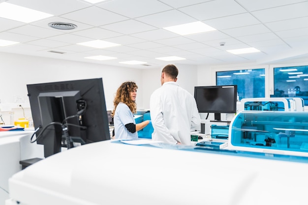 Dos técnicos de laboratorio conversan en medio de máquinas de análisis de sangre en un laboratorio limpio y brillante del hospital