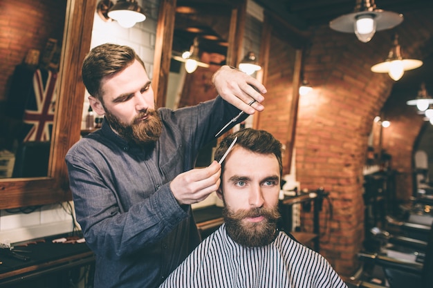 Dos tipos barbudos están en una barbería. El peluquero está cortando el cabello de su cliente con unas tijeras y un cepillo pequeño.