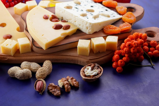 Dos tipos de queso con frutas, bayas, nueces y frutos secos sobre una tabla de madera. Varios tipos de composición de queso con miel y panecillos sobre un fondo azul.