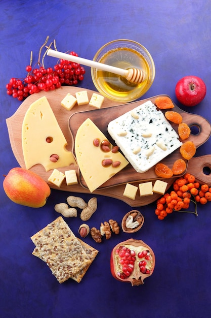 Dos tipos de queso con frutas, bayas, nueces y frutos secos sobre una tabla de madera. Varios tipos de composición de queso con miel y panecillos sobre un fondo azul.