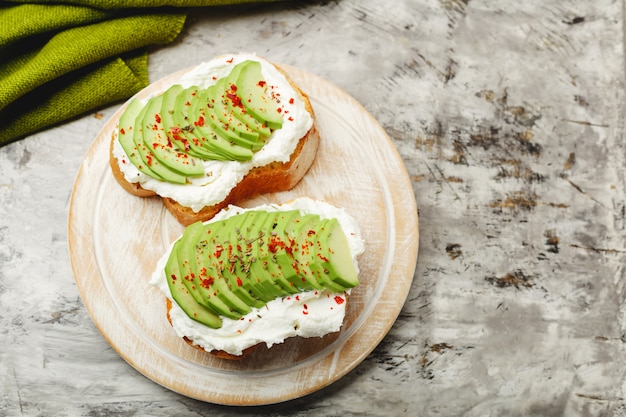 Foto dos tostadas de aguacate, sandwich de aguacate. aguacate fresco en rodajas sobre tostadas de pan de trigo, queso crema. aguacate espolvoreado con chile, especias de albahaca sobre un fondo de hormigón gris con lugar para texto, vista superior