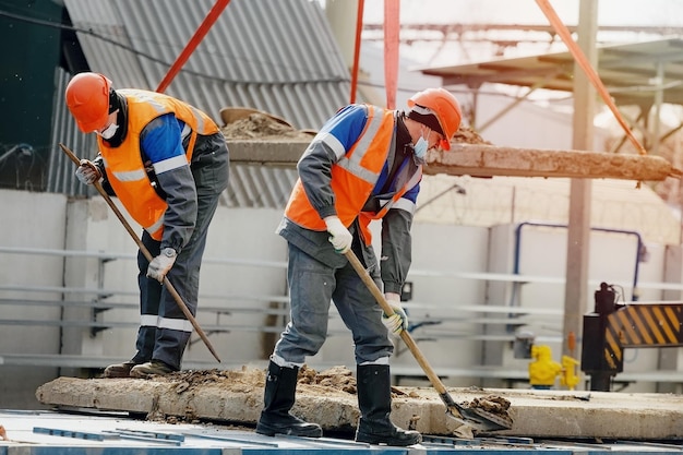 Dos trabajadores con cascos, ropa de trabajo y una máscara médica trabajan con palas en un sitio de construcción Trabajo físico duro