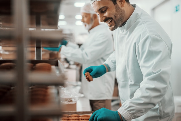 Foto dos trabajadores caucásicos dedicados vestidos con uniformes blancos estériles recogiendo y empacando galletas en cajas. interior de la planta de alimentos.