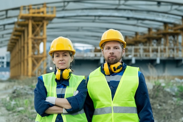 Dos trabajadores de la construcción serios en uniforme y cascos de pie uno al lado del otro