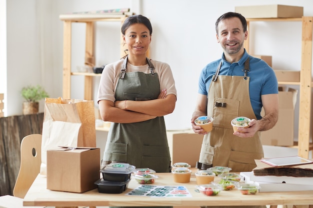 Foto dos trabajadores vistiendo delantales sonriendo mientras envían pedidos en la mesa de madera, servicio de entrega de alimentos